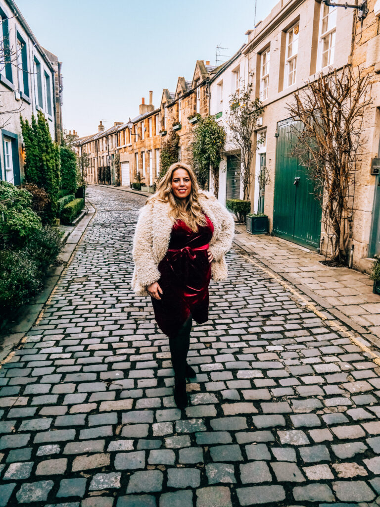 woman in red dress and fur coat standing on cobblestone street in edinburgh scotland